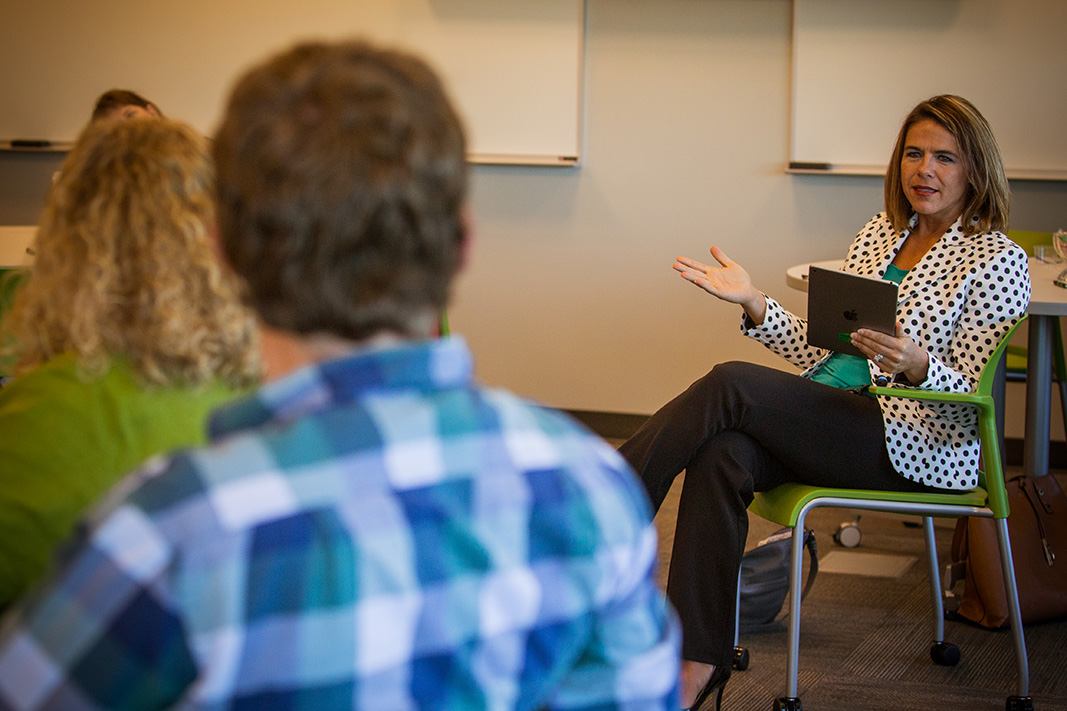 A seated faculty member holds an ipad and motions to students